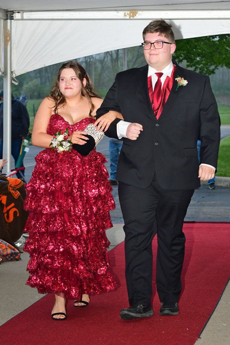 Young couple walking arm-in-arm on a red carpet; woman in a pink sequined gown with a corsage and clutch, man in a black suit with a red vest and boutonniere.