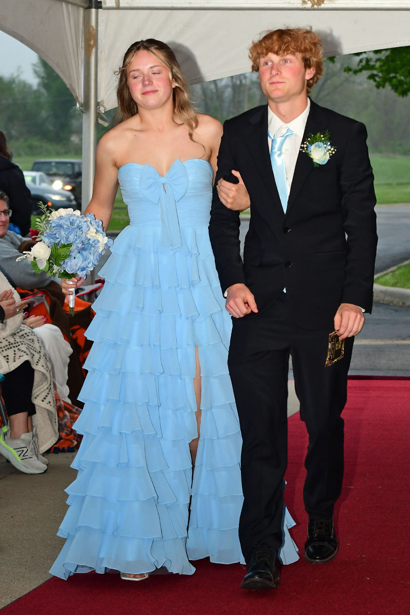 Young couple walking arm-in-arm on a red carpet: girl in a light blue strapless, layered gown with bouquet; guy in a black suit with boutonniere.