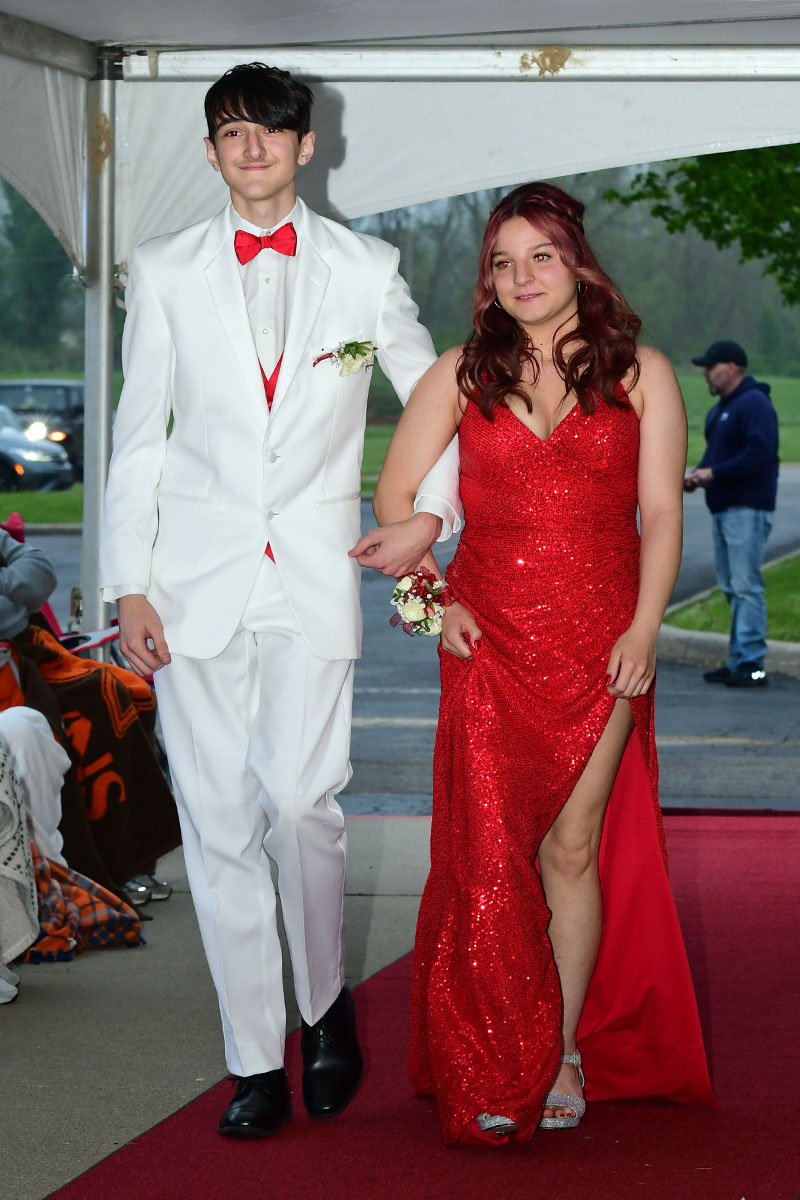 Two teens walk arm-in-arm on a red carpet; boy in a white suit with red bow tie and boutonniere, girl in a red sequined gown with a high slit.