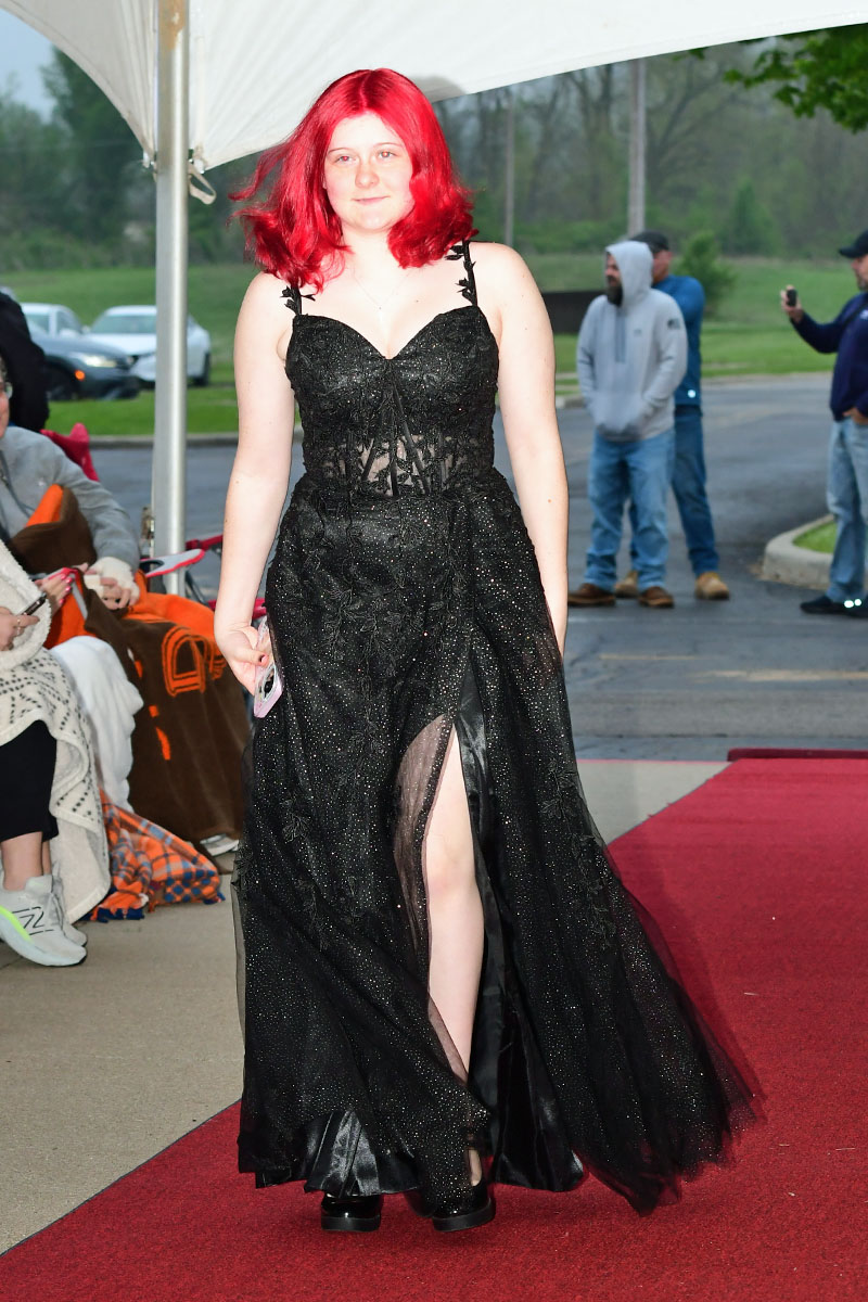 Woman with bright red hair in a glittery black evening gown walking the red carpet outdoors.