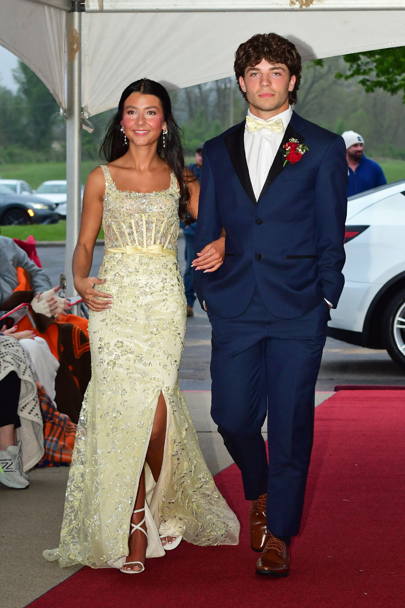 Young couple on a red carpet: woman in a cream beaded gown and heels, man in a navy suit with a cream bow tie, walking arm in arm.