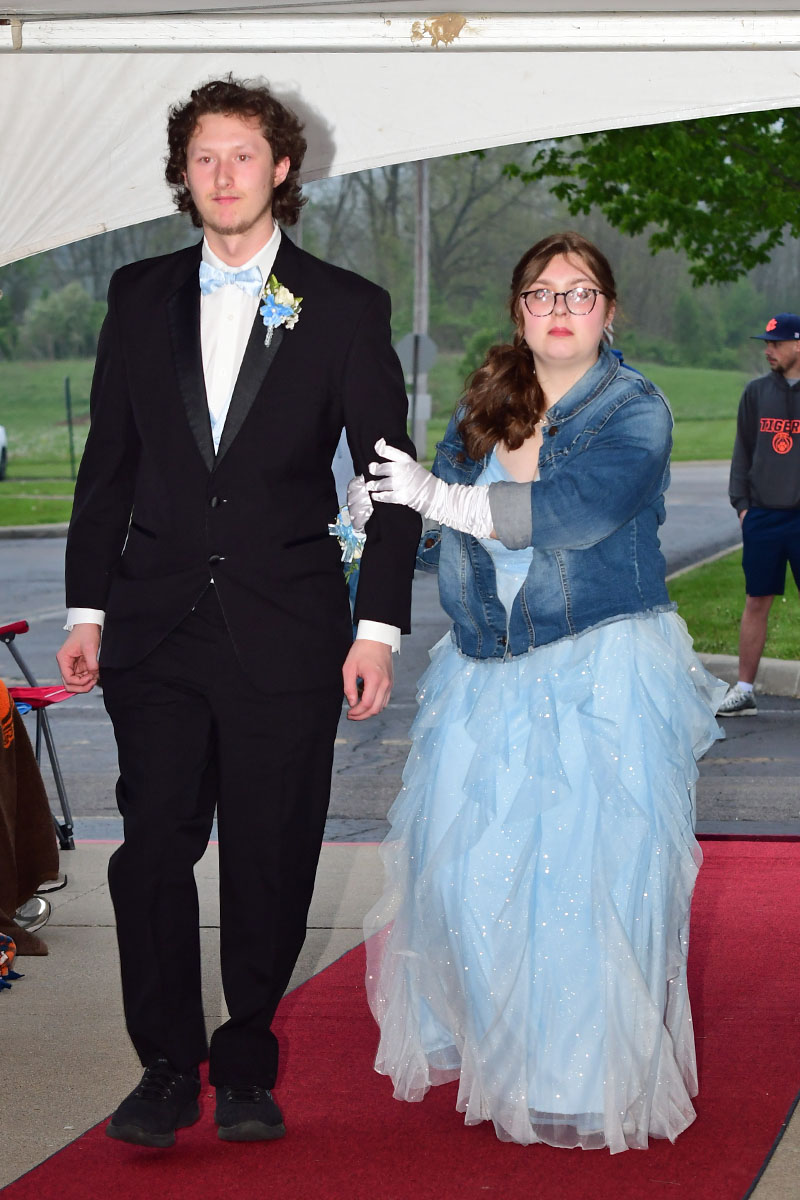 Couple walking on a red carpet at a formal event; man in black tuxedo with light blue bow tie and corsage, woman in a pale blue gown and denim jacket.