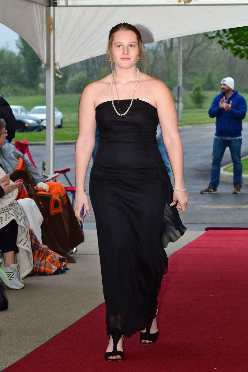 Woman in a black strapless gown walks the red carpet at an outdoor fashion event with spectators nearby, pearl necklace in view
