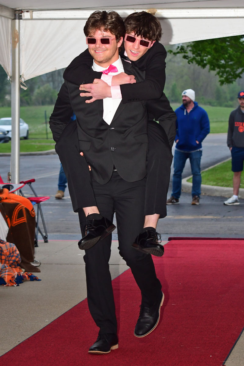 Two men in suits walk on a red carpet; one carries the other piggyback while both wear pink sunglasses outdoors under a tent.