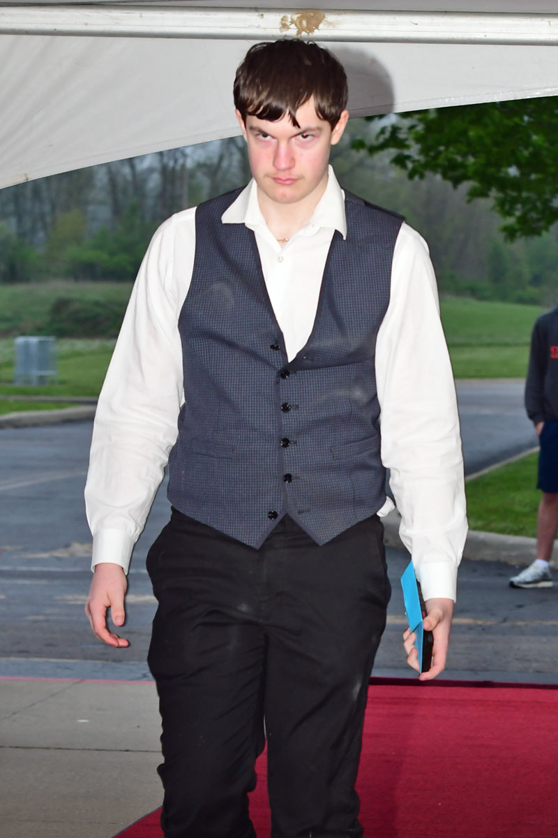 Young man in a white shirt and dark vest walking on a red carpet outdoors, holding a blue folder.