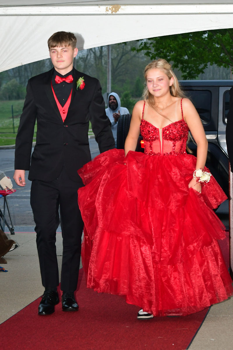 Young couple arriving on a red carpet: a boy in a black suit with red vest and bow tie and a girl in a bright red ball gown with a corsage on her wrist.
