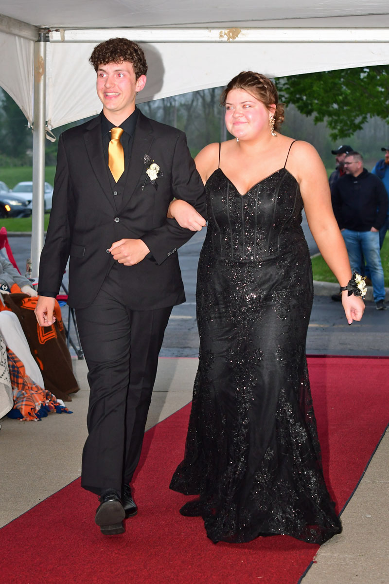Two teens walking arm-in-arm down a red carpet, boy in black suit with gold tie and boutonniere, girl in black sequined gown with corsage on wrist.