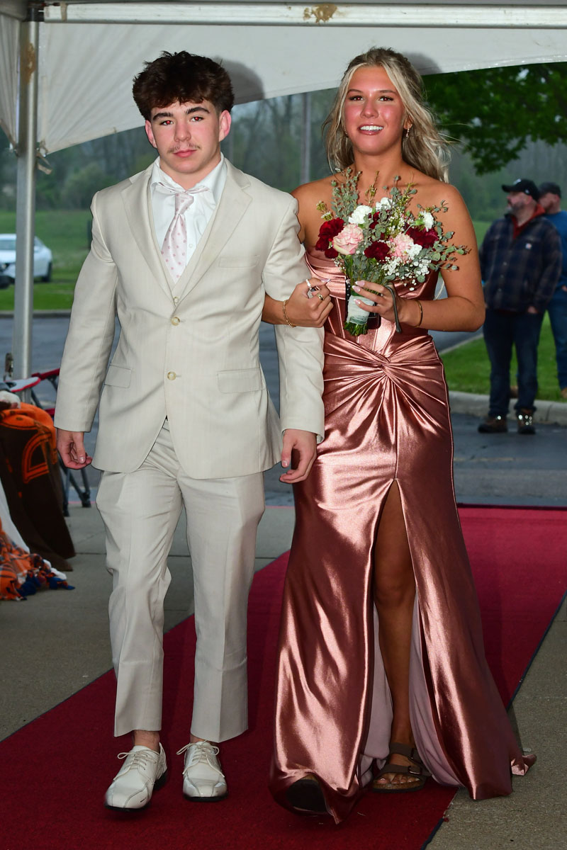 Couple walking arm in arm on a red carpet: man in a beige suit, white shirt and tie, and woman in a rose metallic gown holding a bouquet of pink and red flowers.