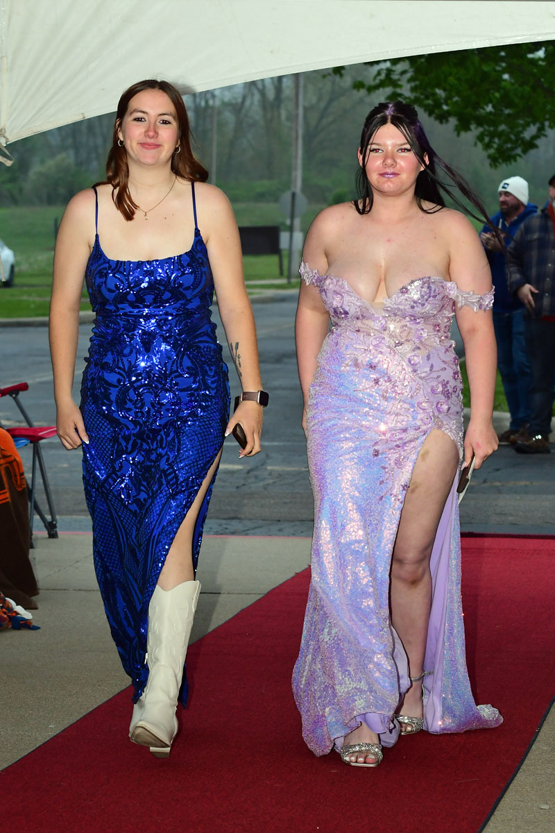 Two young women walk the red carpet in formal gowns: one in a blue sequin dress with a thigh slit and white boots, the other in a lavender off‑shoulder gown with a high slit and glittery sandals.
