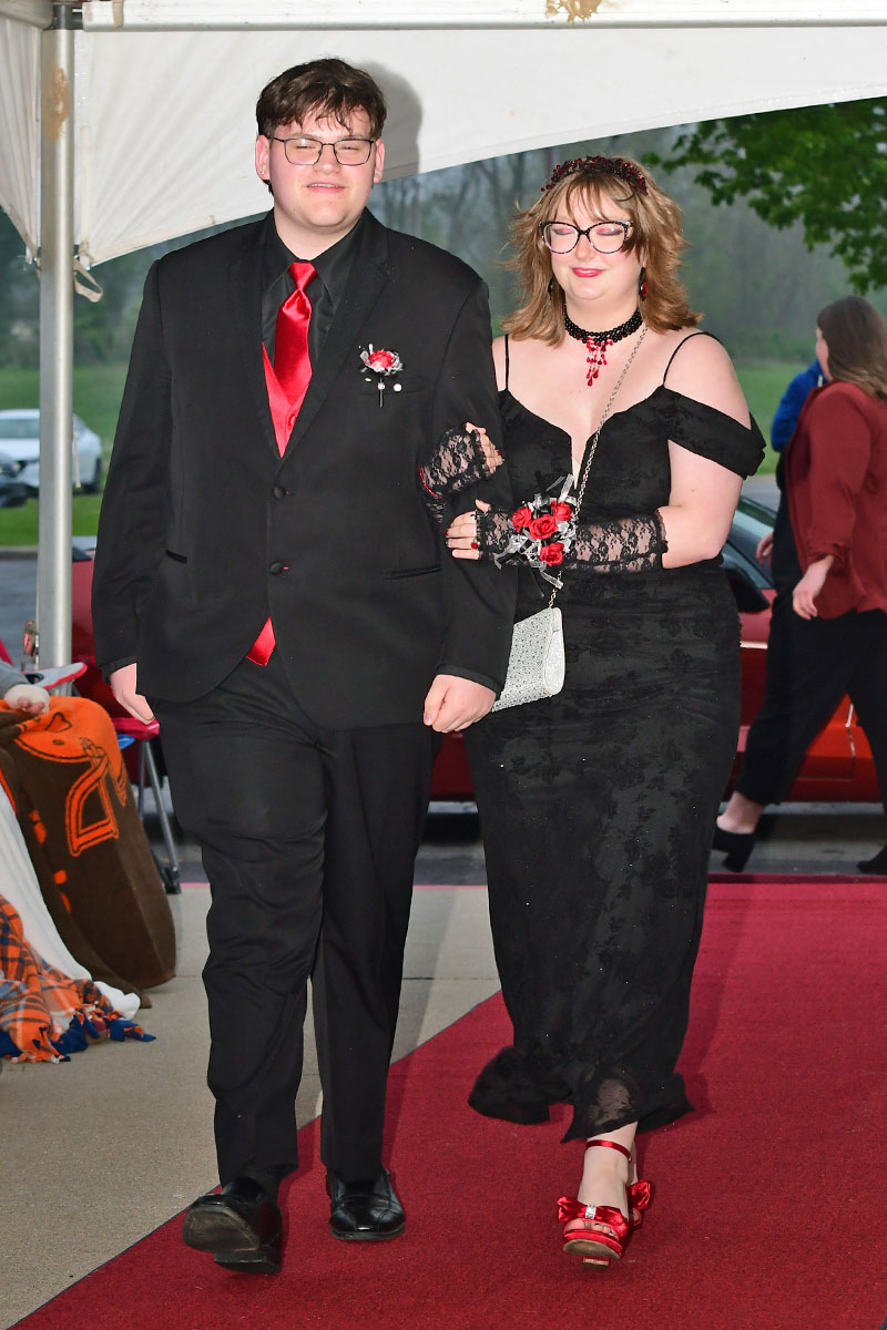 Couple walking down a red carpet at a formal event; man in a black suit with a red tie and boutonniere, woman in a black lace gown with red accessories and gloves.