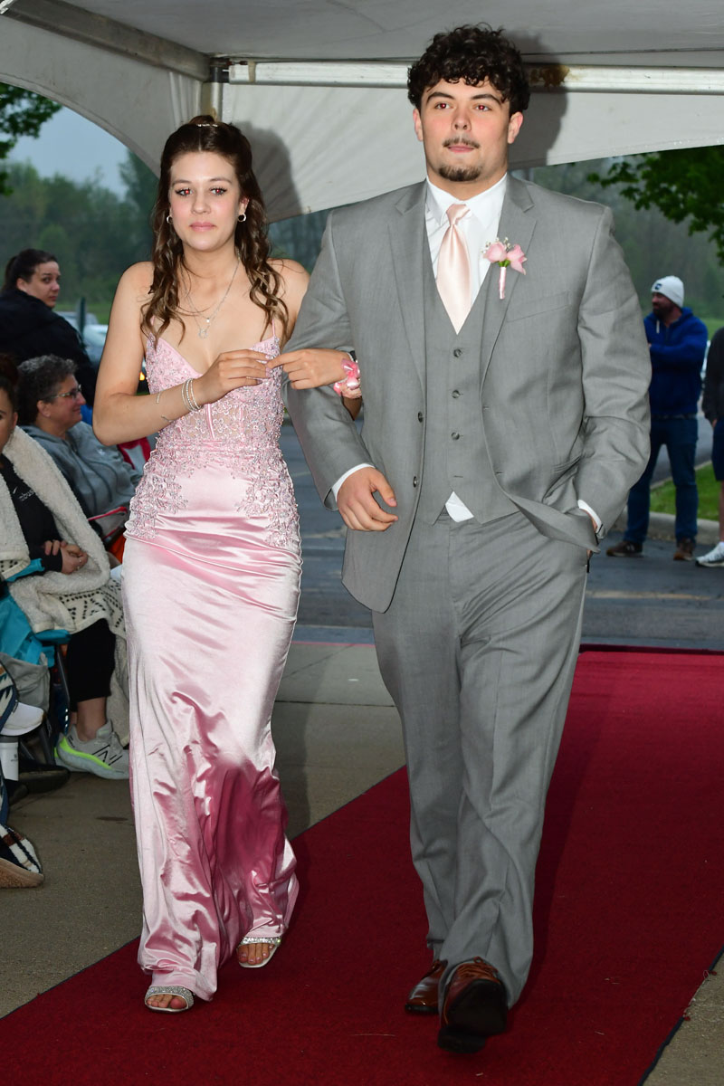 Young couple walks arm-in-arm down a red carpet; girl in a pink beaded gown, boy in a gray suit with pink boutonniere.