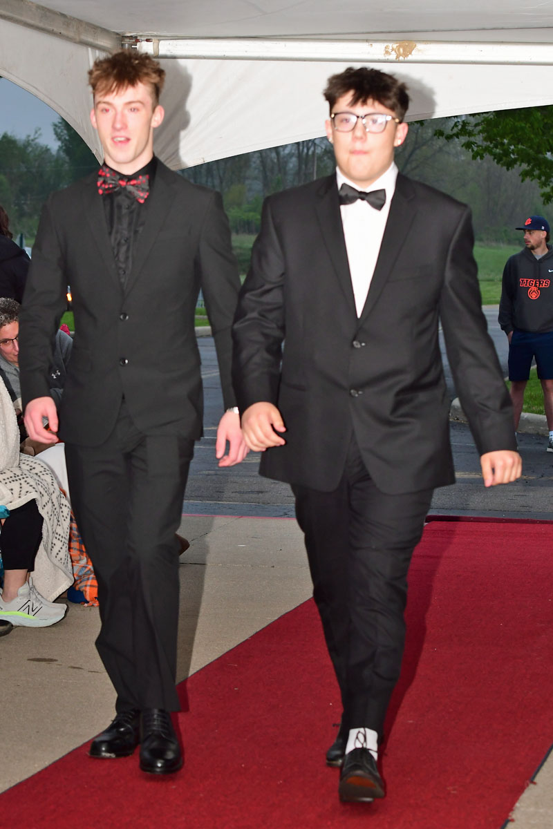 Two young men in black tuxedos walk down a red carpet under a white tent at an outdoor event, one wearing a red bow tie and the other a black bow tie and glasses.