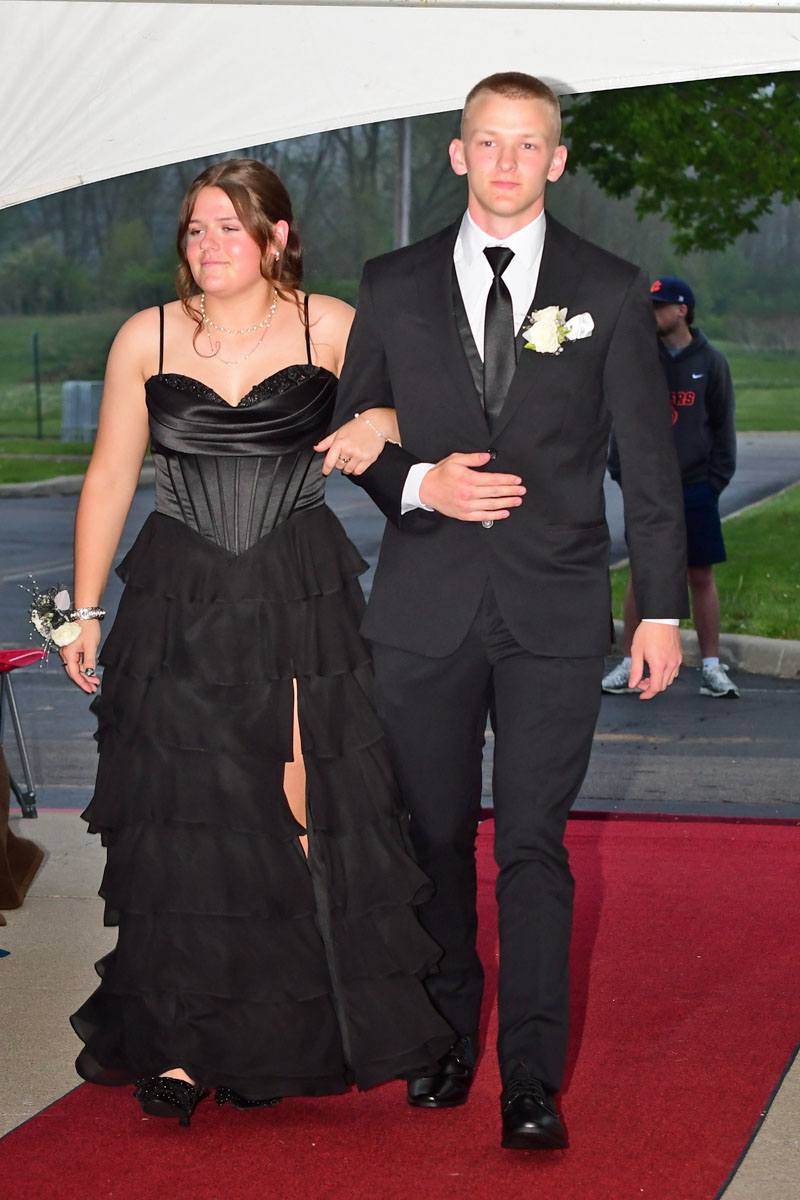 Couple walking arm-in-arm down a red carpet at a formal event; woman in a black tiered gown and man in a black suit with white shirt and tie, boutonniere on his lapel.