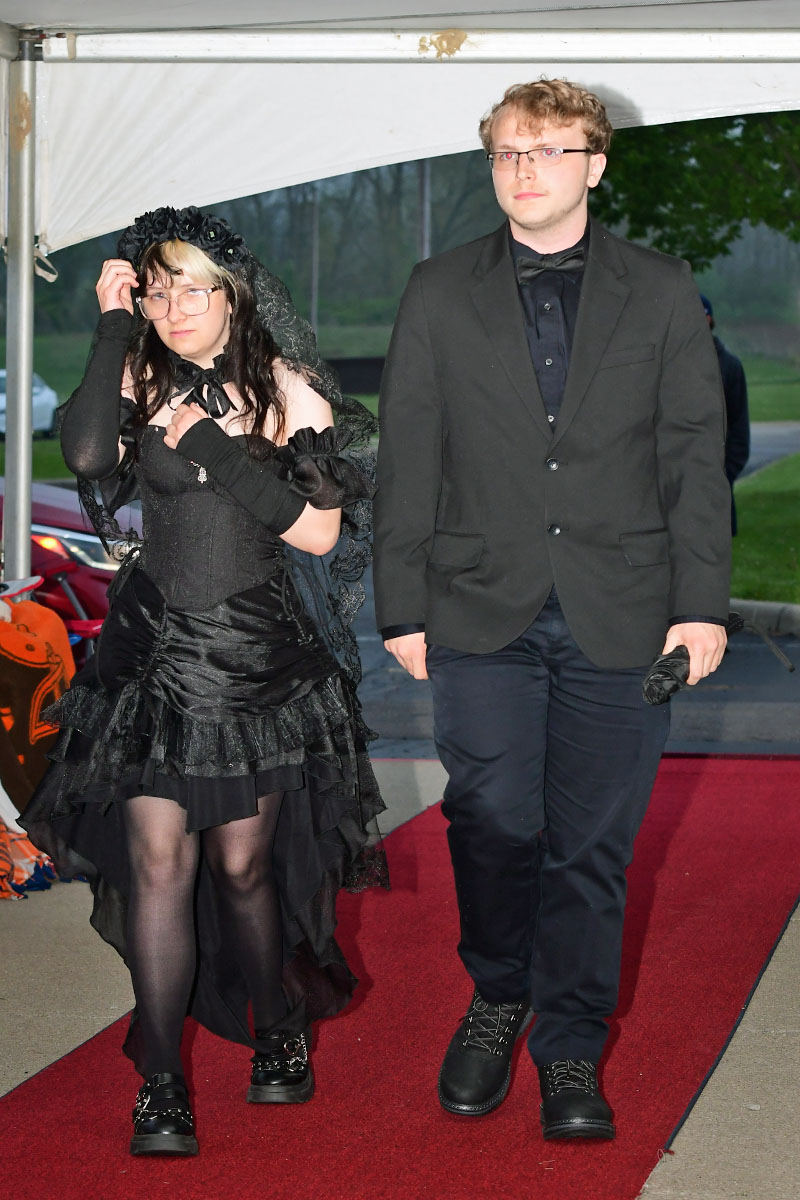 Two people walk the red carpet under a white tent: a woman in a black Victorian-style dress with lace veil and glasses, and a man in a black suit with a bow tie.