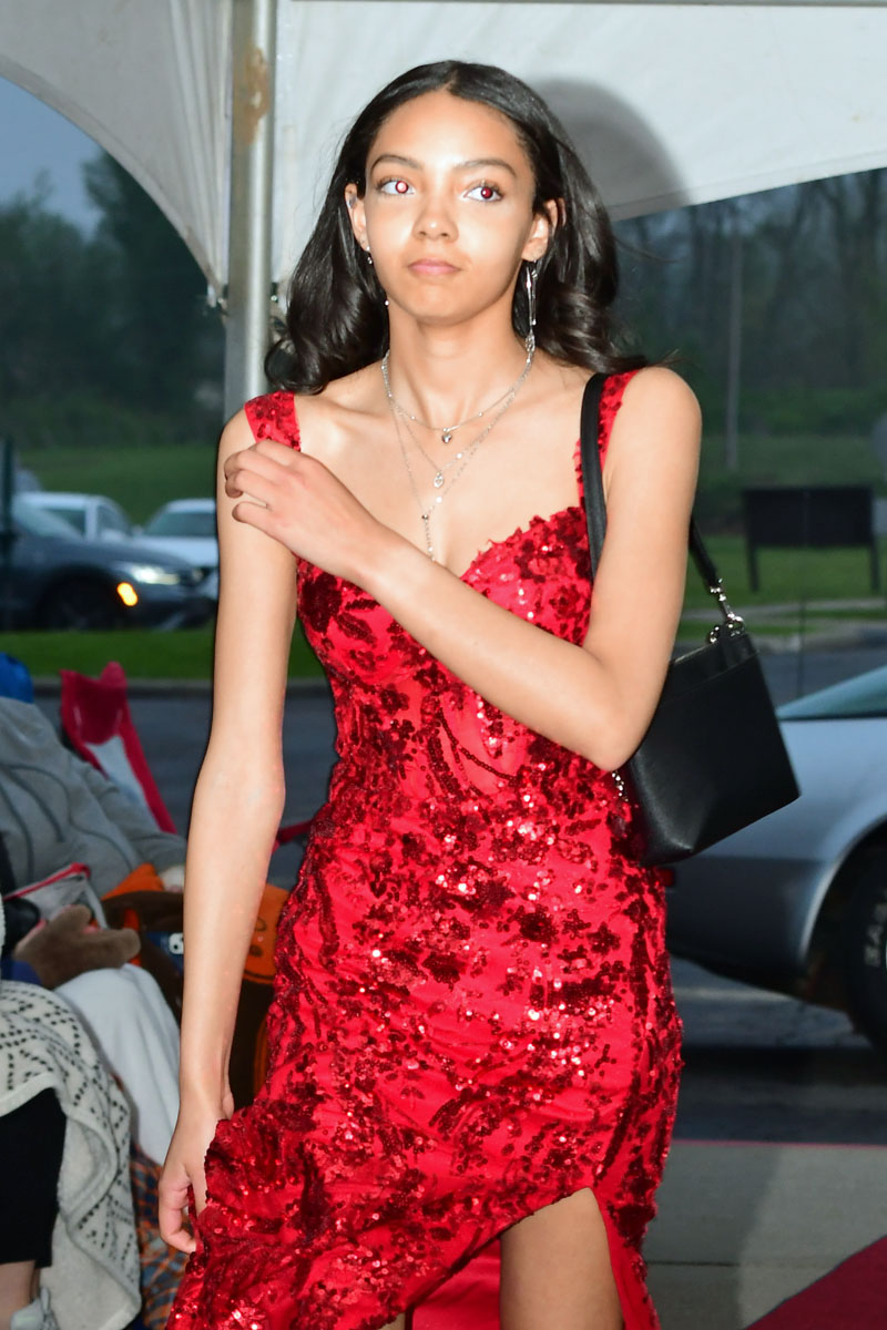 Young woman in a red sequined gown walking on a red carpet under a white tent, carrying a black shoulder bag.