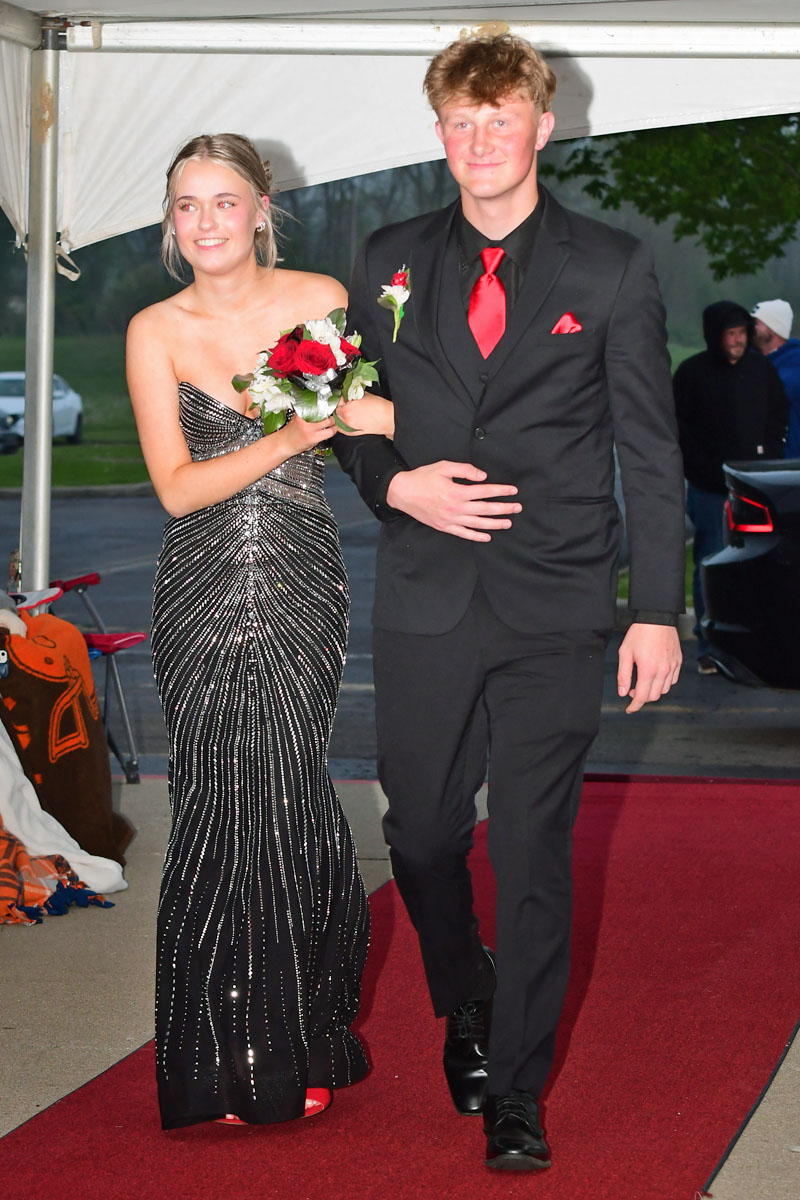Teen couple in formal wear walking the red carpet: girl in a silver sequined gown holding a rose bouquet, boy in a black suit with a red tie and boutonniere.