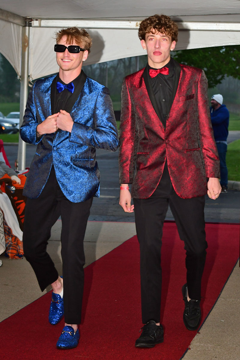 Two young men walk a red carpet wearing shiny patterned suits—blue on the left with a blue bow tie and sunglasses, red on the right with a red bow tie.