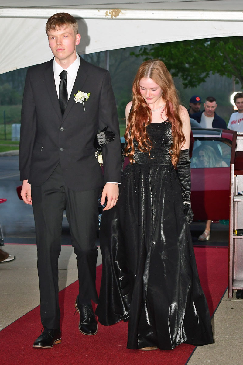 Young man in a black tuxedo with boutonniere walks beside a woman in a shimmering black gown on a red carpet under a tent.