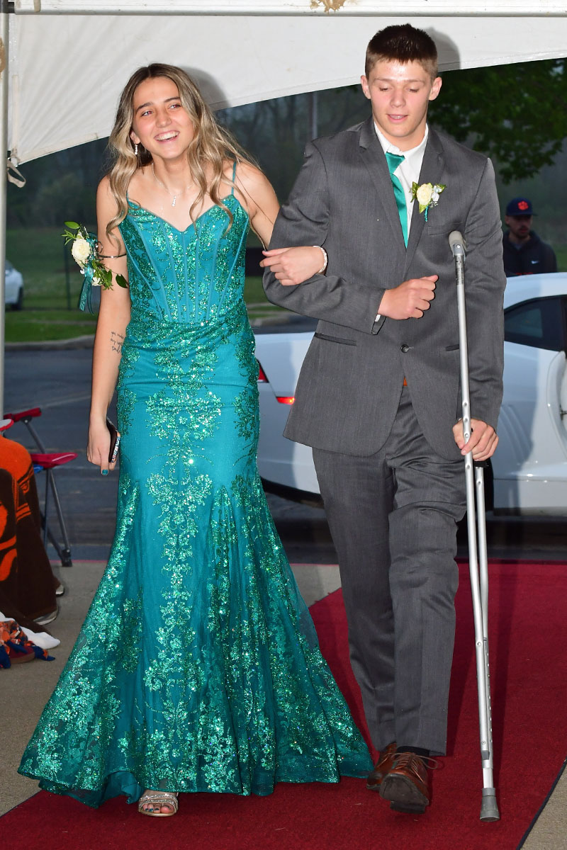 Teen couple on a red carpet; girl in a teal sequined prom dress, boy in a gray suit with a crutch.