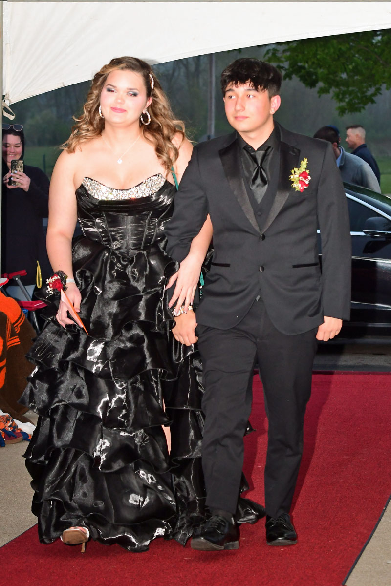 Couple walking the red carpet at a formal event; woman in a black tiered gown, man in a black suit with boutonniere.