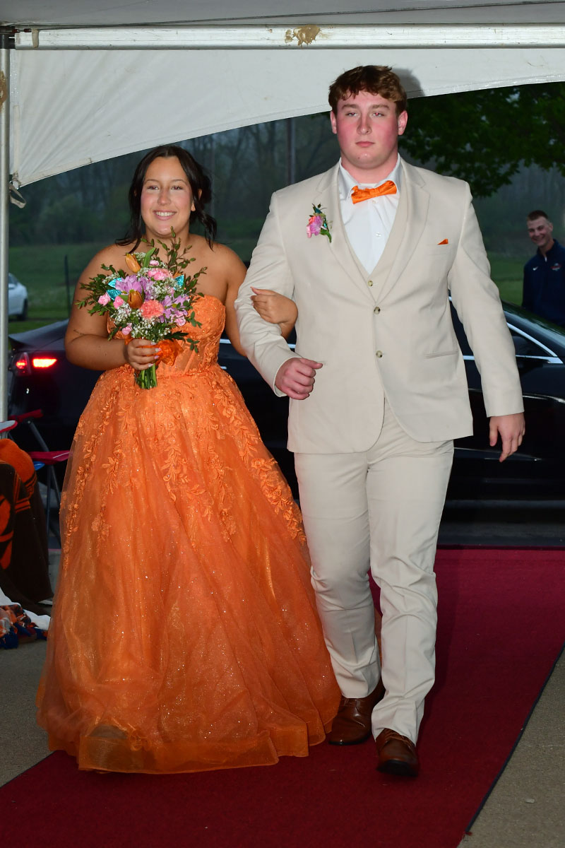 Teen couple walking arm-in-arm down a red carpet: girl in an orange strapless ball gown holding a bouquet, boy in a beige suit with an orange bow tie.