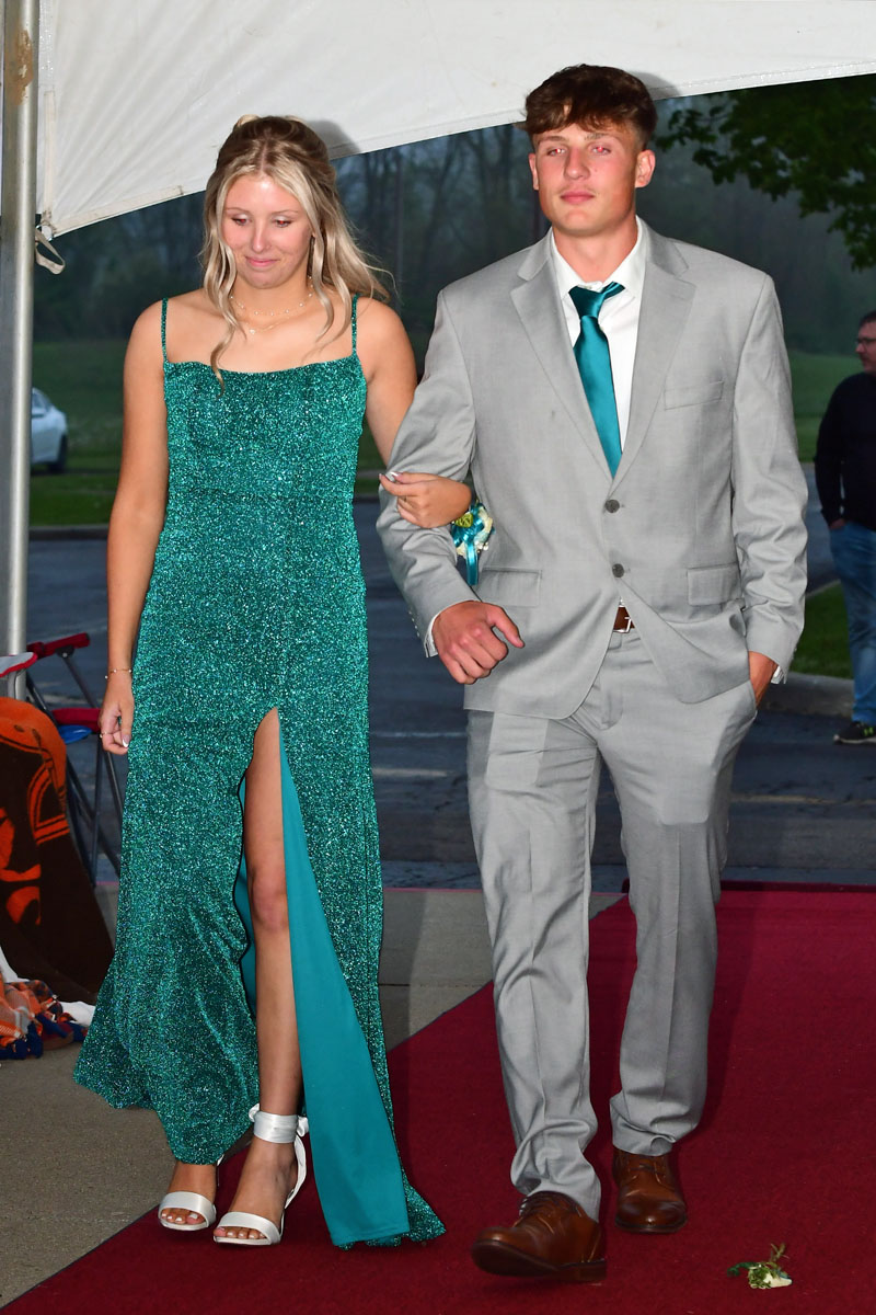 Couple walking arm-in-arm on a red carpet; woman in a teal glitter dress, man in a gray suit and teal tie outside under a tent.