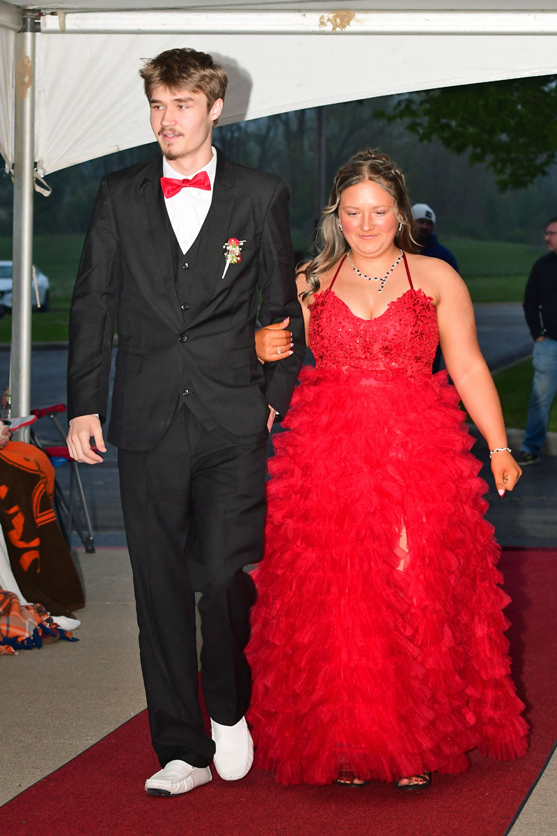 Couple walking arm-in-arm on a red carpet; man in a black suit with red bow tie and boutonniere, woman in a bright red ballgown.