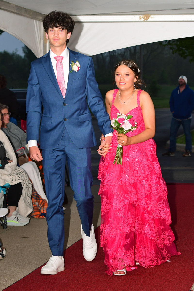 Teens at prom walking arm in arm on a red carpet; boy in a blue suit with pink tie and boutonniere, girl in a pink lace dress holding a bouquet.