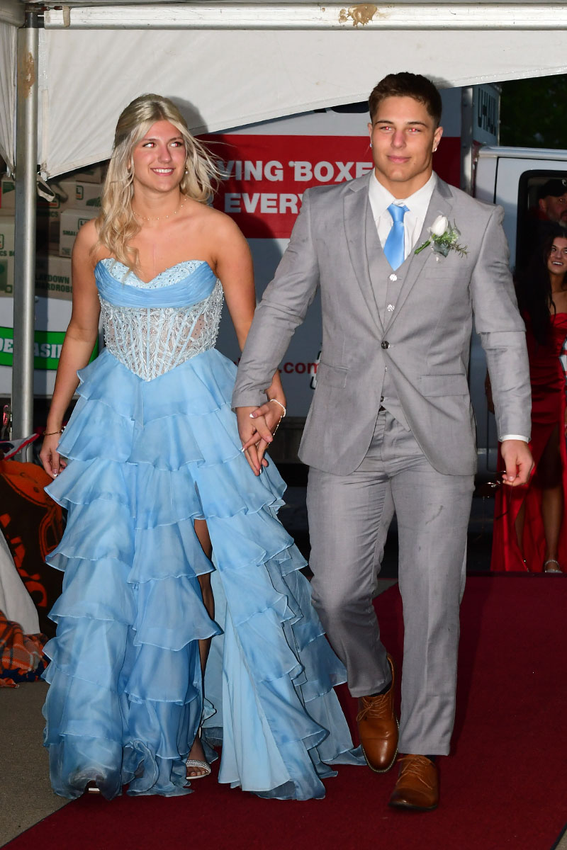 Young couple walking hand in hand on a red carpet; woman wears a light blue layered ball gown, man in a grey suit with a blue tie and boutonniere.