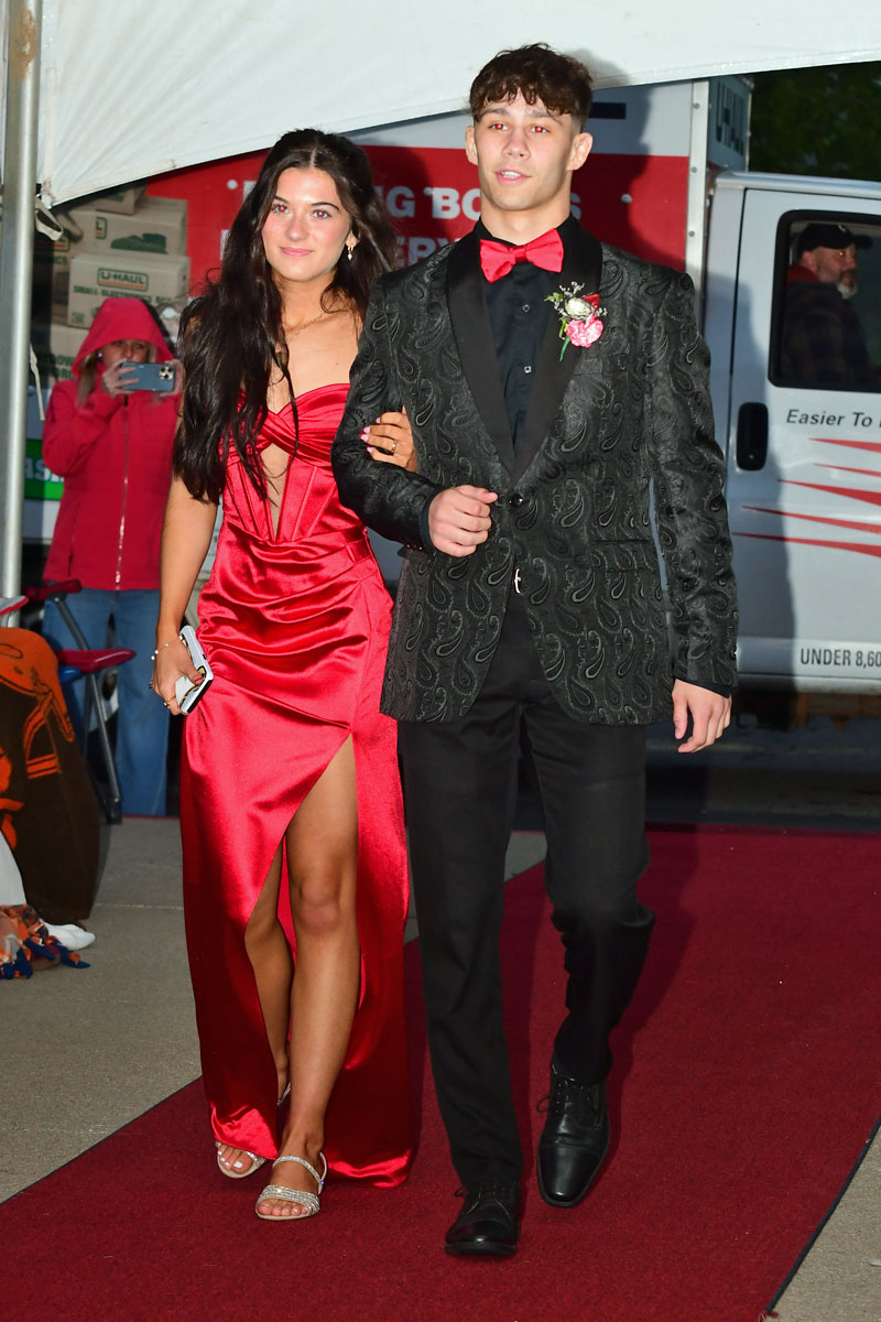 Couple walking the red carpet: woman in a red satin gown with a high slit and silver sandals, man in a black patterned blazer with a red bow tie.