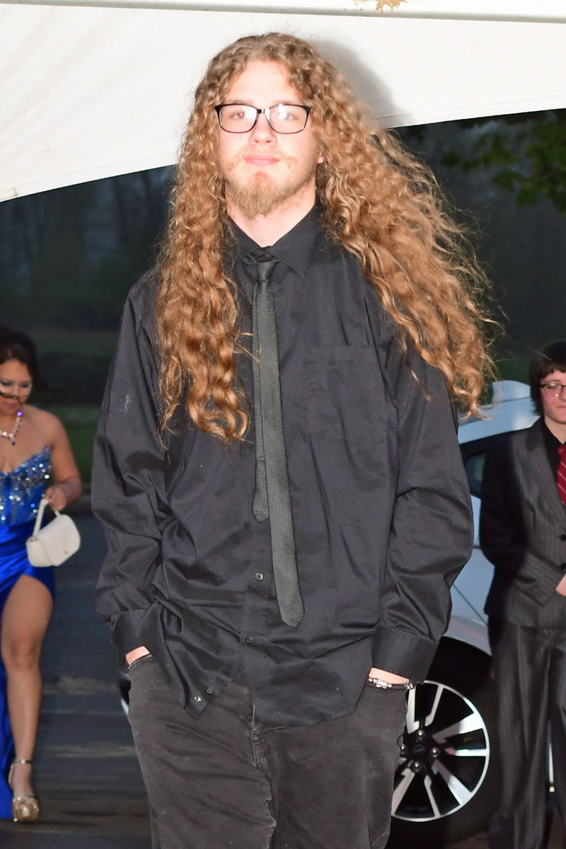 Man with long curly hair and glasses in a black shirt and tie at an outdoor formal event.