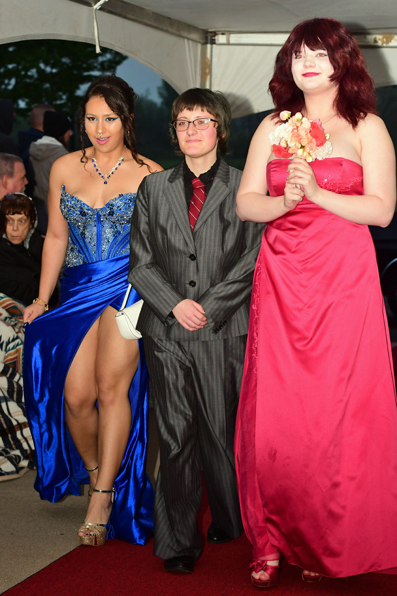 Three young people posing on a red carpet: a blue-gowned woman, a person in a gray pinstripe suit, and a pink-dressed woman holding a bouquet.