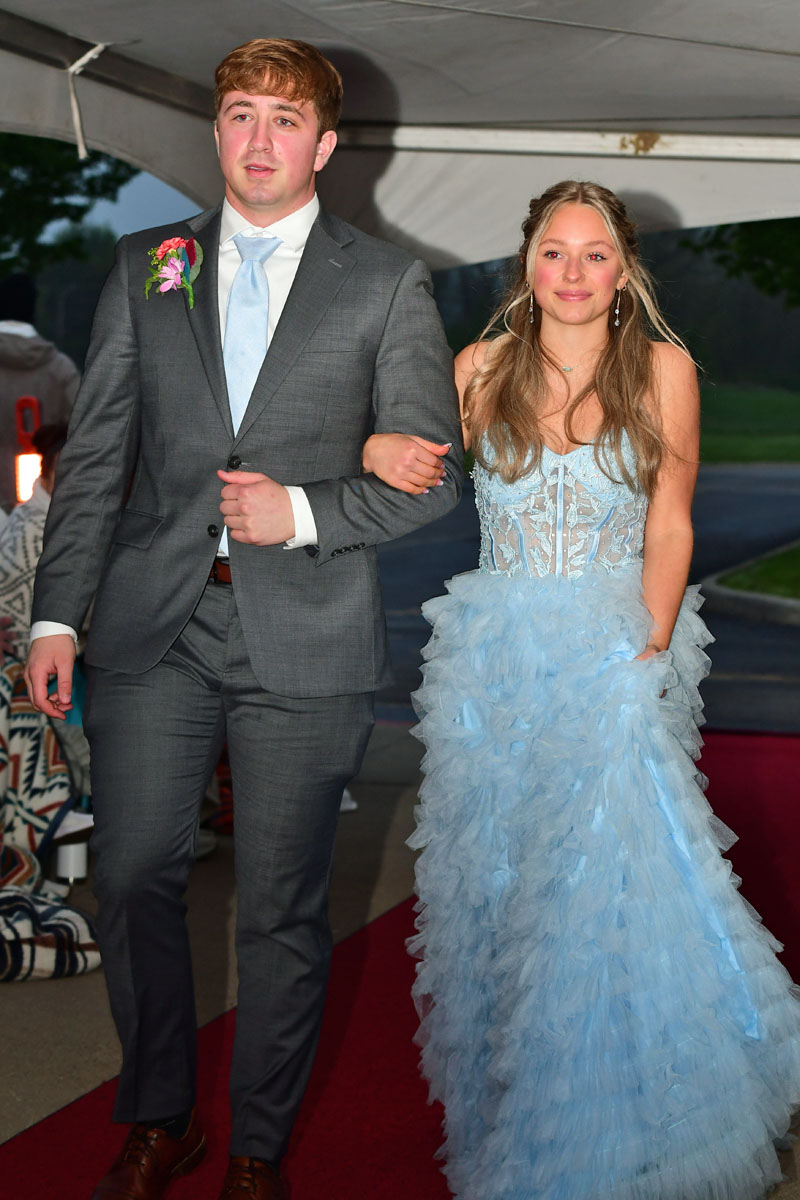 Teen couple walking arm-in-arm on a red carpet; the boy in a gray suit with a boutonniere and light blue tie, the girl in a light blue lace and tulle gown smiling.