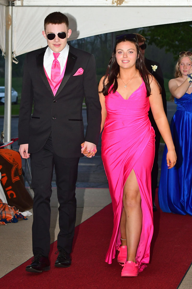 Young couple walking hand in hand on a red carpet; man in a black suit with pink tie and sunglasses, woman in a bright pink dress and matching shoes.