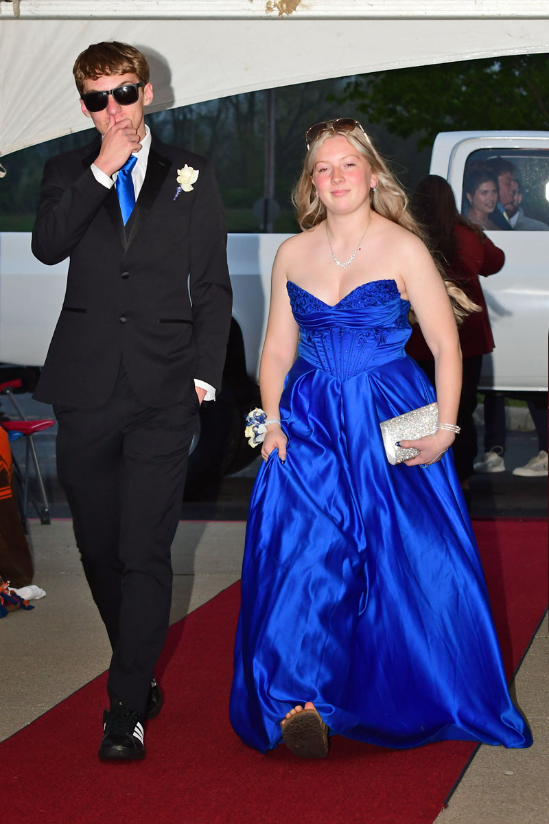 Couple walking the red carpet: young man in a black suit with blue tie and sunglasses, woman in a cobalt blue formal gown and silver clutch.