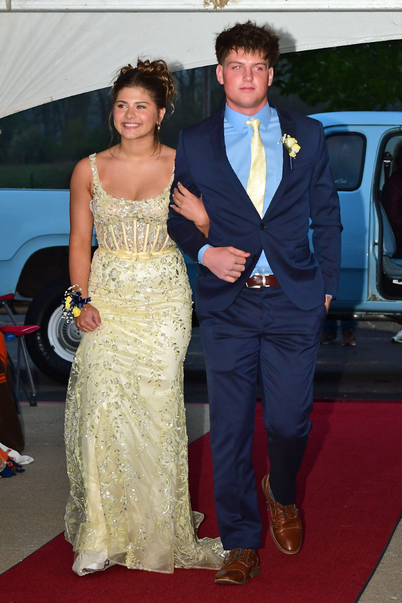 Two teens walk arm-in-arm down a red carpet at prom: a girl in a cream beaded gown and corsage on her wrist, and a boy in a navy suit with a yellow tie next to her.