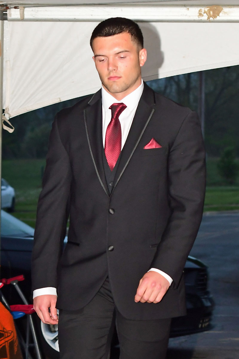 Man in a black suit with a red tie and pocket square walking outdoors under a tent or canopy.