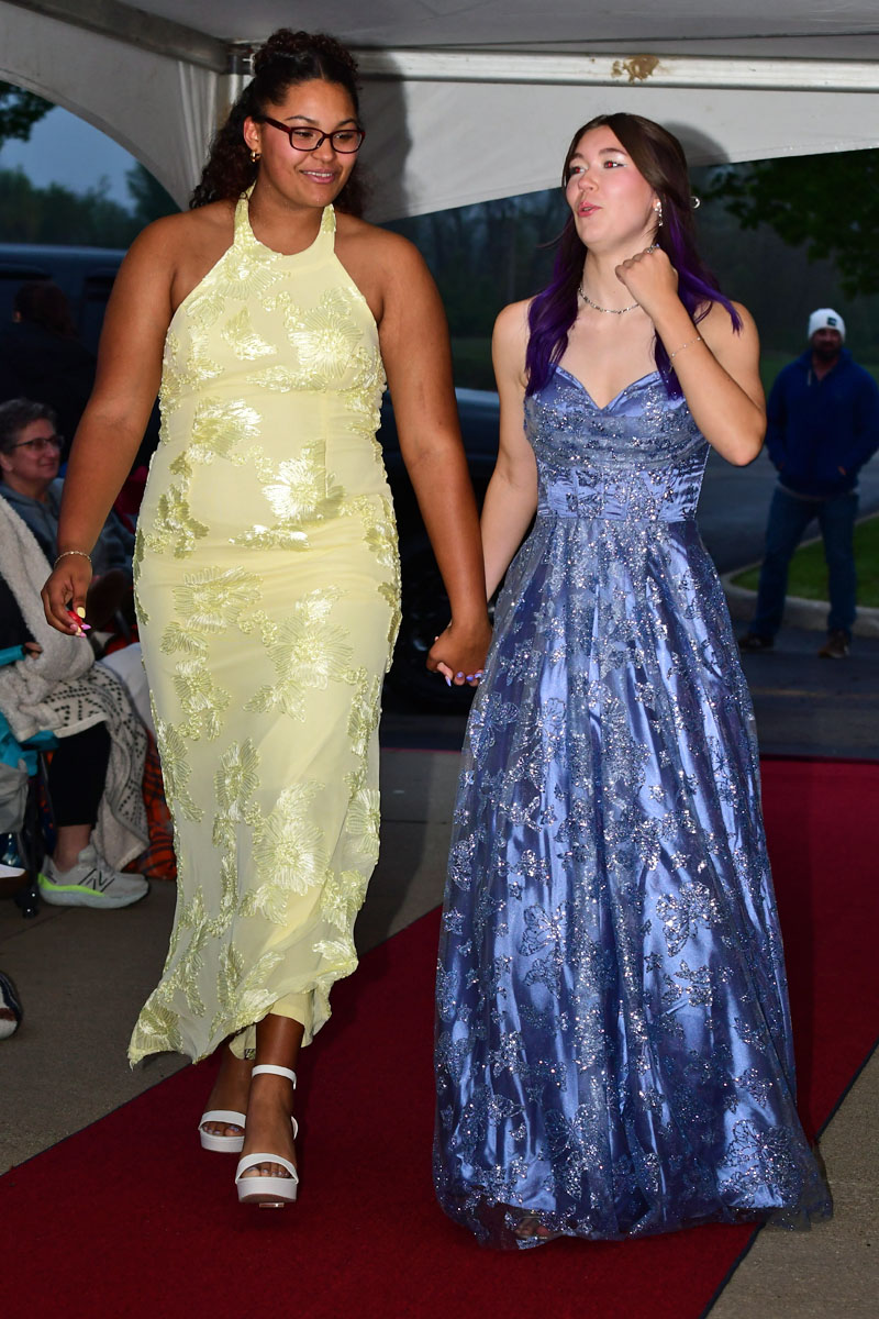 Two young women in formal gowns walk hand in hand on a red carpet under a canopy, smiling.