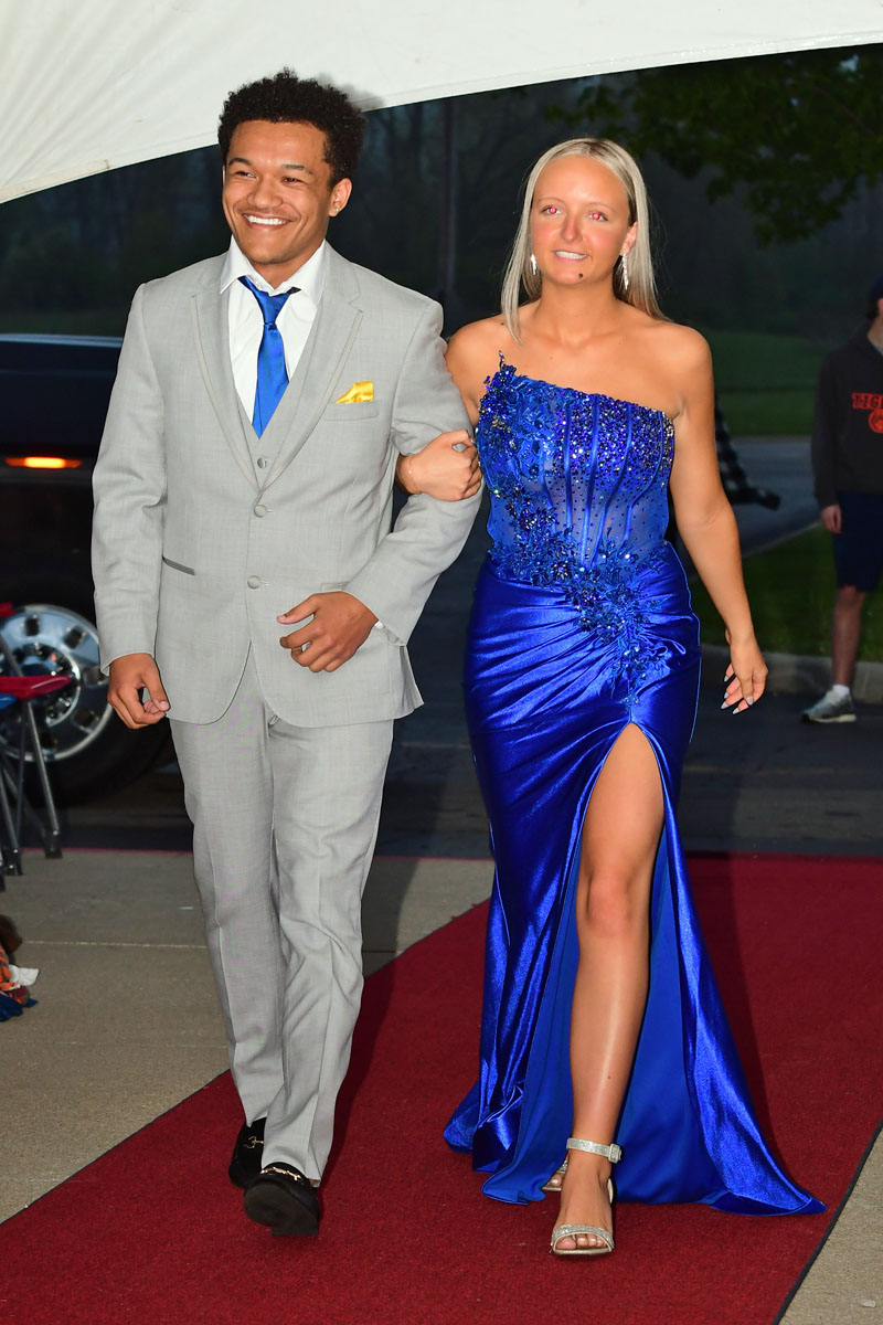 Man in a light gray suit with blue tie escorts a woman in a sparkling royal blue gown along a red carpet, smiling side by side with arms linked.