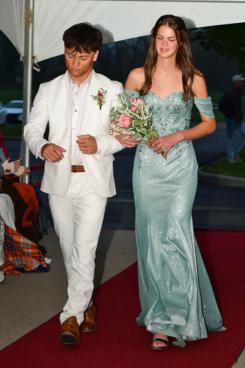 Teenage couple walking a red carpet; the boy wears a white suit and brown shoes, the girl in a glittery light-blue gown holding a pink bouquet.
