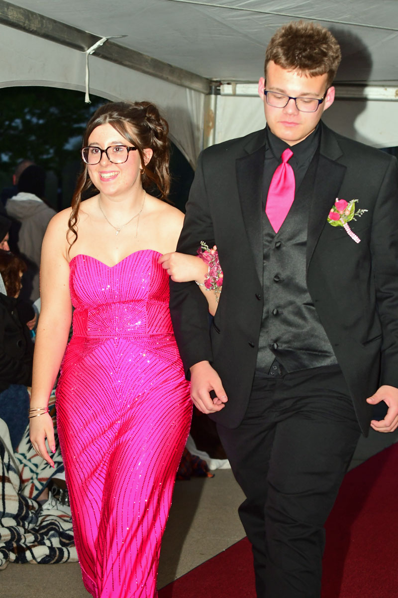 Young couple walking down a red carpet at a formal event; girl in a pink sequined strapless dress and glasses, boy in a black suit with a pink tie and boutonniere.