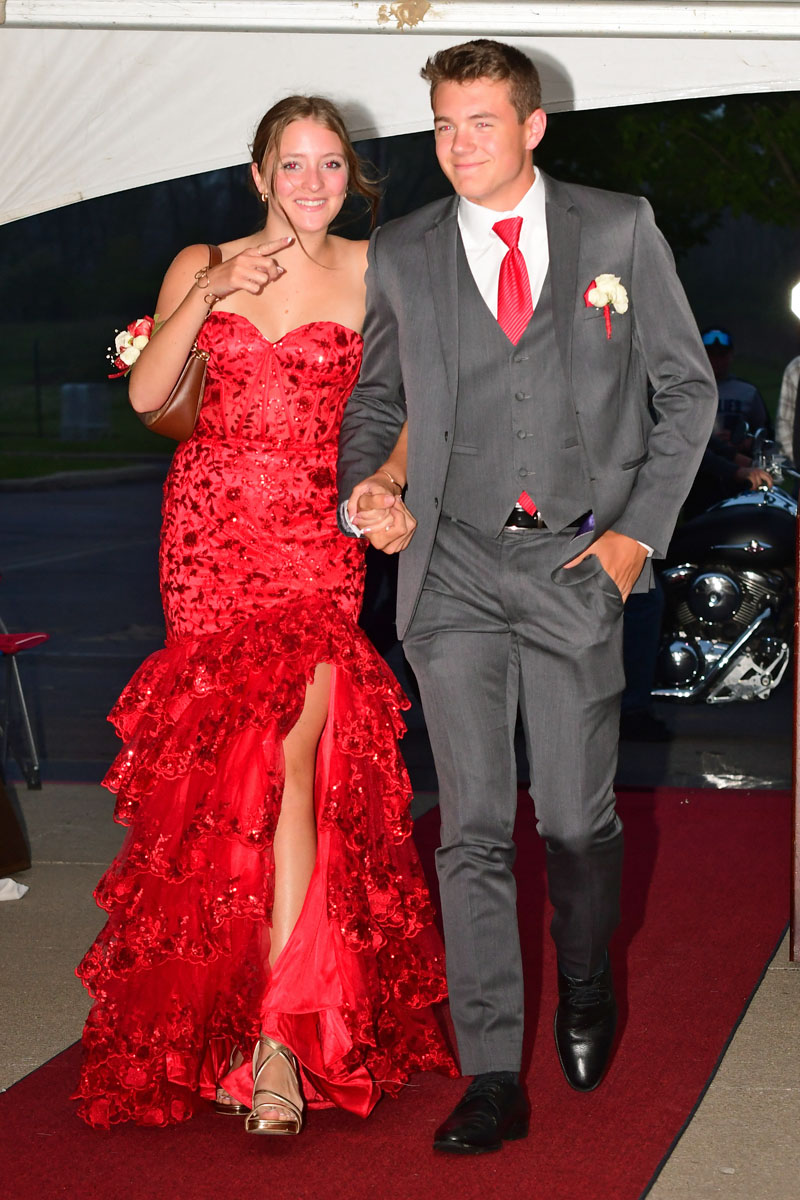Couple walking the red carpet: girl in a bright red ruffled gown and the boy in a gray suit with a red tie, holding hands.