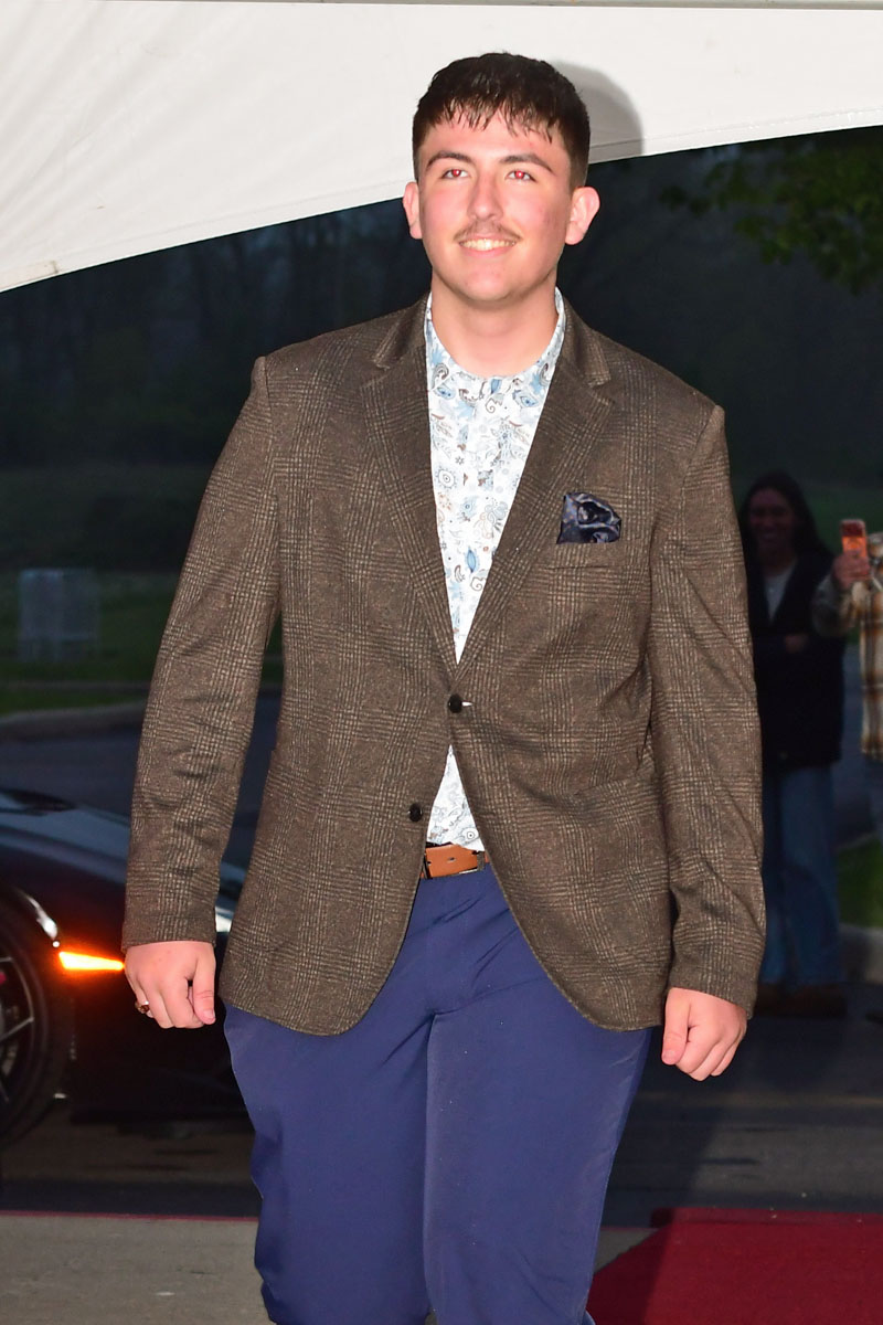 Young man in a brown plaid blazer, patterned shirt, and blue pants posing on a red carpet. Pocket square visible in blazer pocket.