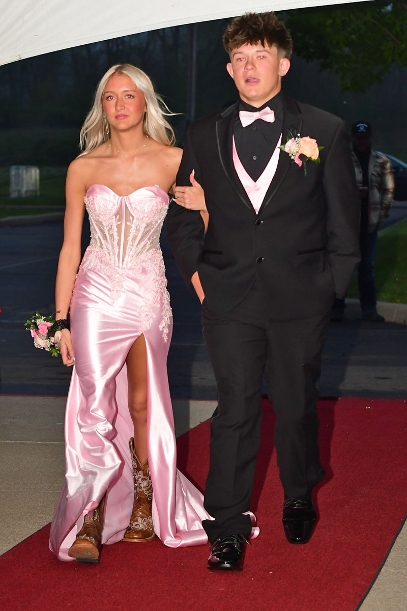 Prom night couple walking the red carpet: woman in a pink corseted gown with a high slit and boots, man in a black suit with pink vest and bow tie.