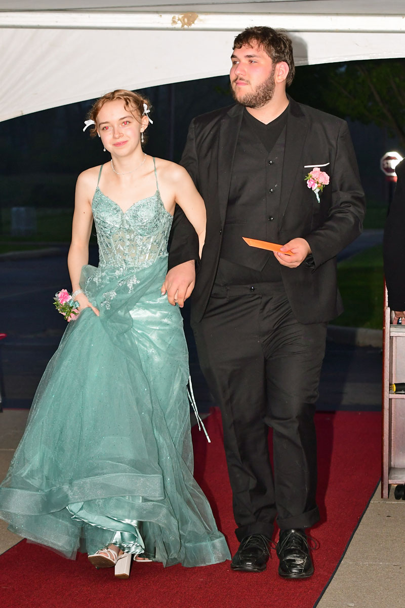 Couple walking a red carpet; woman in teal gown and floral hair accessories, man in black suit with boutonniere and envelope in hand.