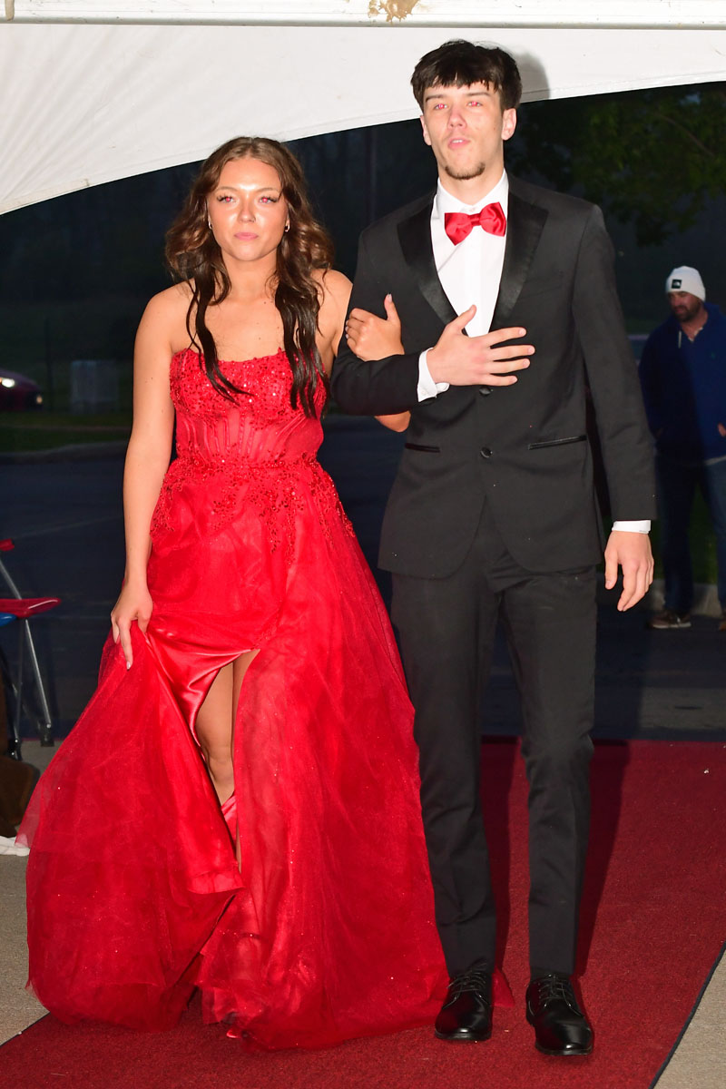Couple walking arm-in-arm on a red carpet: girl in a sparkling red evening gown and boy in a black tuxedo with a red bow tie.