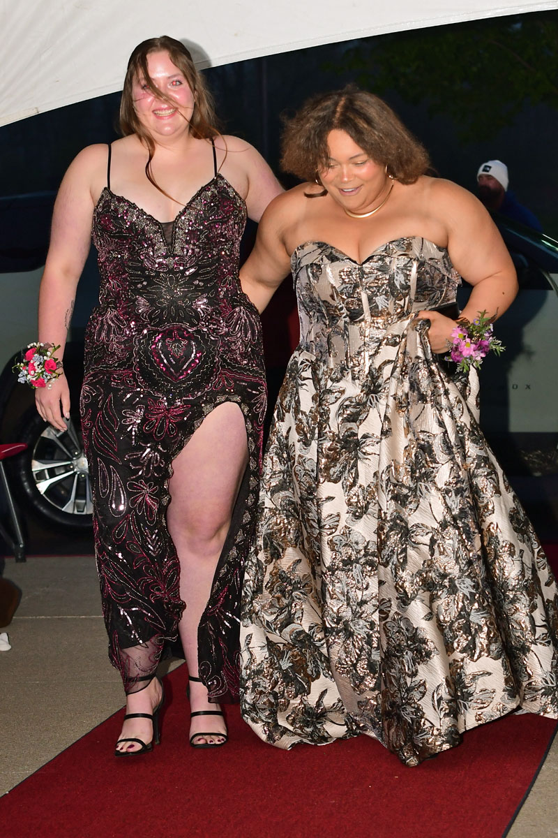 Two women in elegant sequined gowns walk arm in arm on a red carpet under a white canopy, smiling.