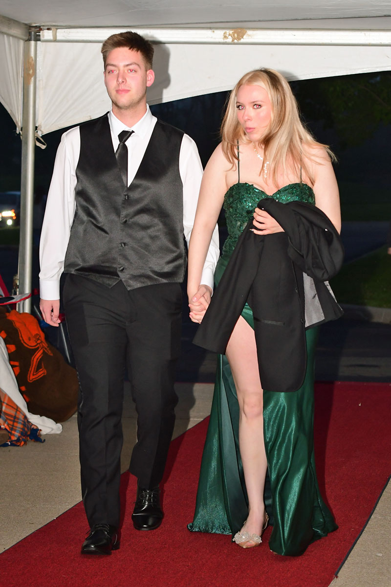 Couple walking on a red carpet at a formal event; man in a black vest and tie, woman in an emerald green gown holding a jacket