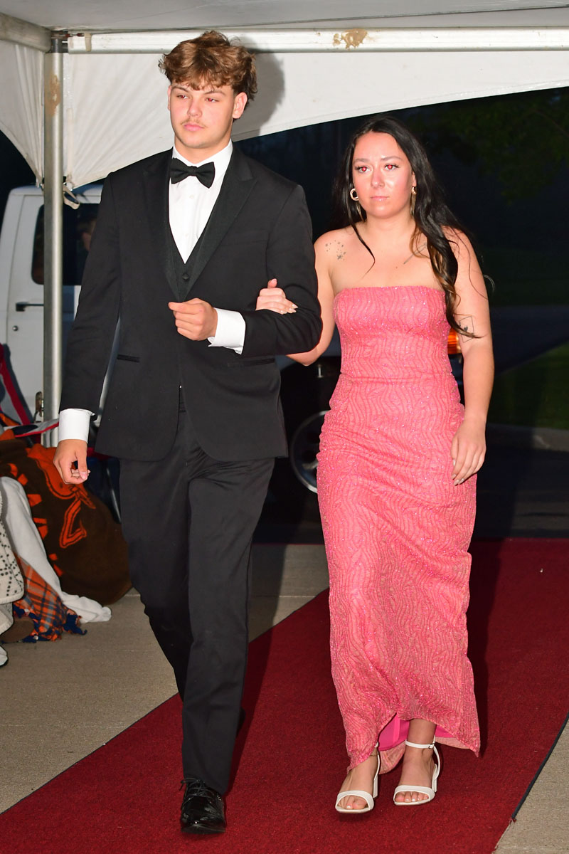 Couple walking a red carpet: man in black tuxedo assisting a woman in a pink strapless gown under a white canopy at night.