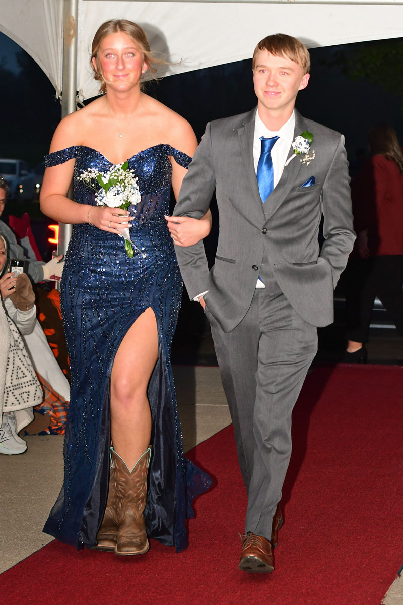 Couple walking arm-in-arm down a red carpet at a formal event: woman in a sparkly navy off-shoulder gown with a high slit and brown cowboy boots, man in a gray suit with a blue tie and boutonniere.